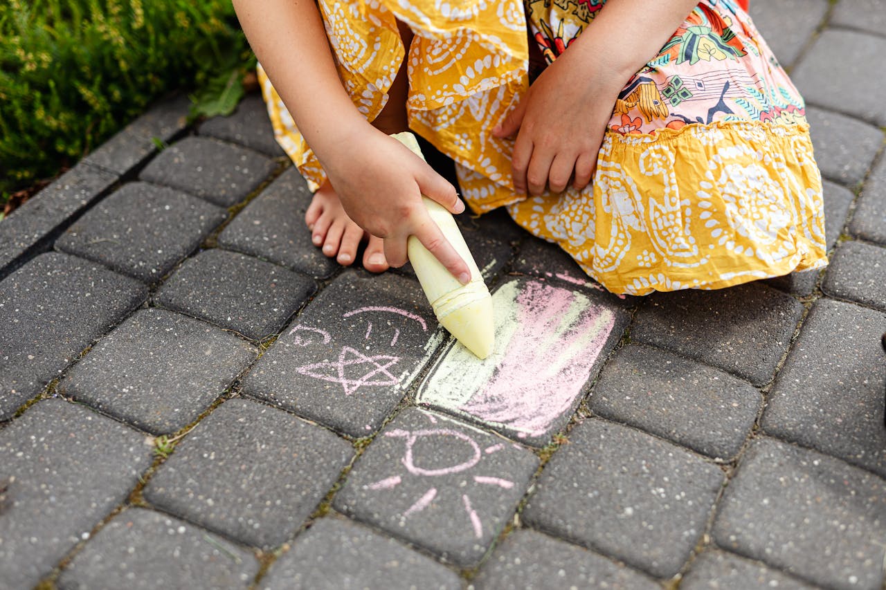 Home A child in a colorful dress draws with chalk on a paved walkway, creating playful doodles.